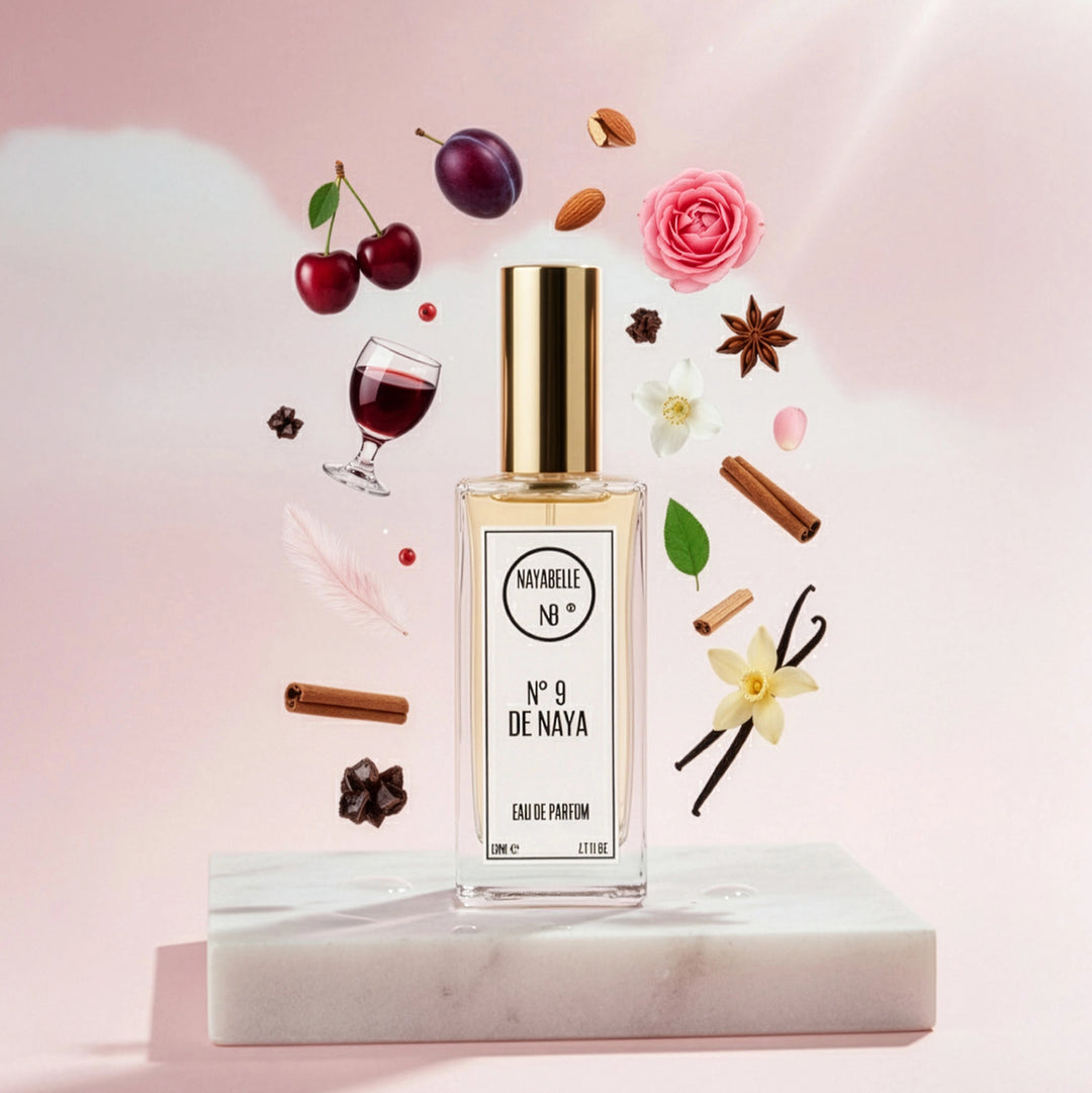 Parfum N° 9 de Naya par Nayabelle. Flacon posé sur un socle de marbre blanc, entouré des ingrédients de la fragrance flottant dans les airs : cerise, prune, amande, cannelle, vanille et épices. Photo créative pour e-commerce.