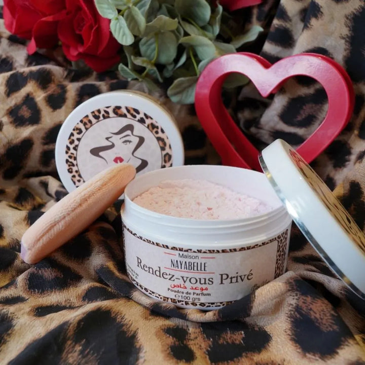 Boîte ouverte de poudre parfumée "Rendez-vous Privé" de Maison Nayabelle, révélant une poudre rose délicate avec une houpette douce posée à côté. Le tout est disposé sur un tissu imprimé léopard, accompagné de roses rouges, d'un cœur rouge en décoration et du couvercle orné d'une silhouette féminine stylisée, créant une atmosphère luxueuse et romantique.